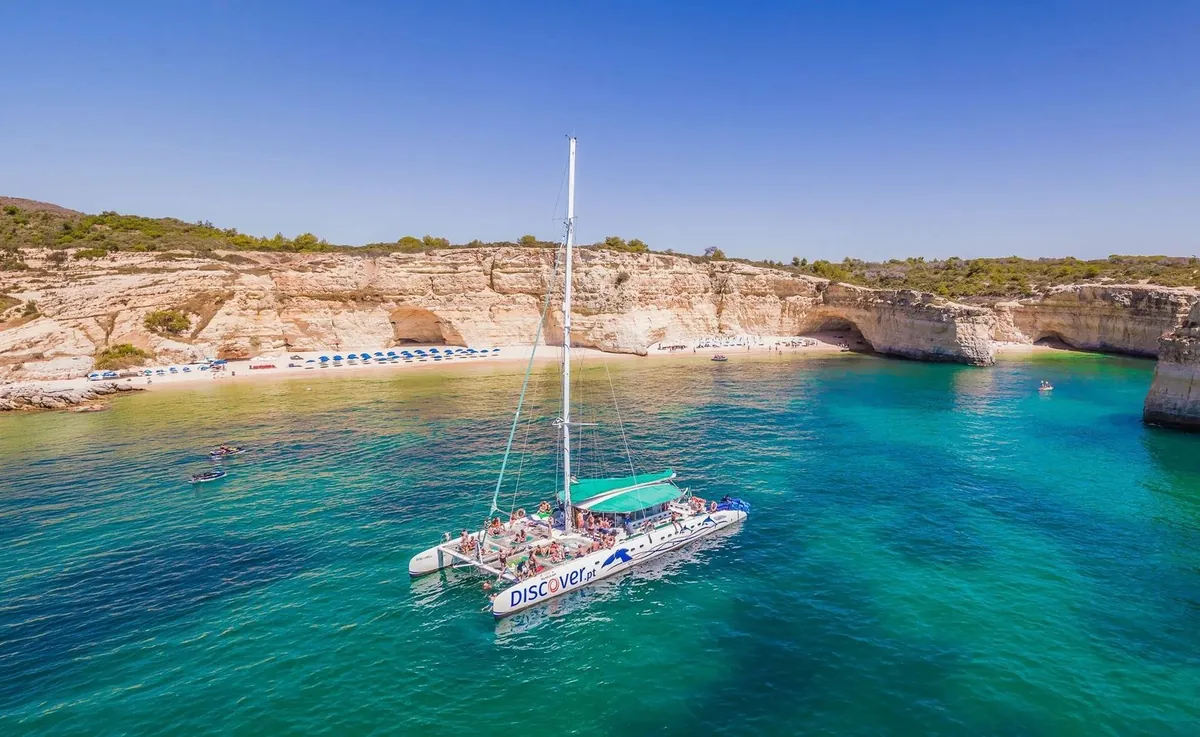 Melhor do Mar sailing catamaran with guests aboard beside the Algarve cliffs.