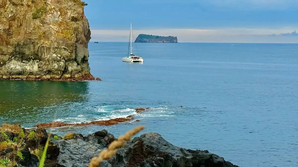 A white SailZen catamaran in clear blue water beside the São Miguel coastline.